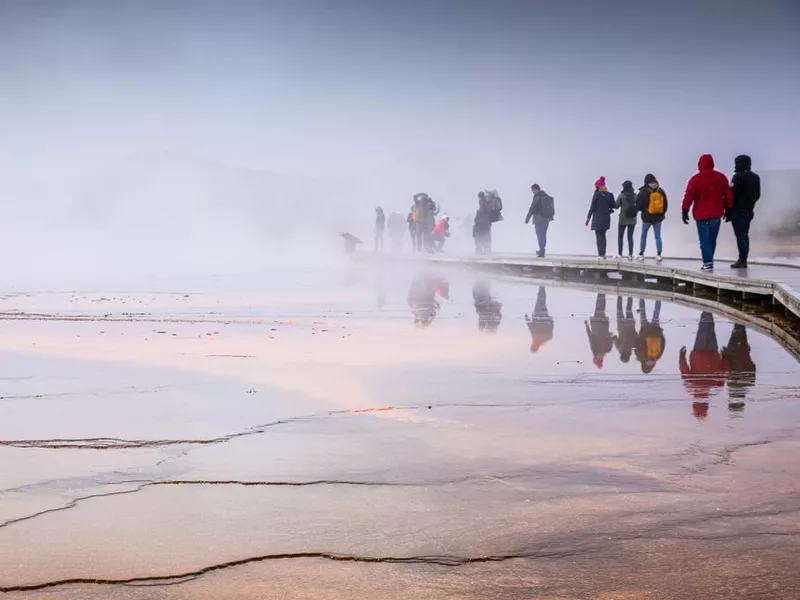 Misty scene of walking tourists on wooden boardwalk inside Grand Prismatic Spring.